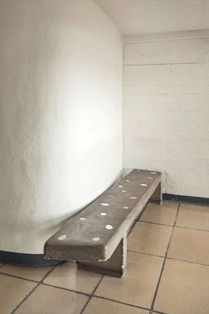 Concrete bench inlaid with small round tiles sits against a curved white textured wall above a terracotta tile floor.