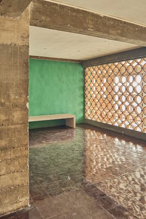 Modernist interior with raw concrete beams, a green wall, built-in bench, polished marble floor and a circular perforated screen letting in 