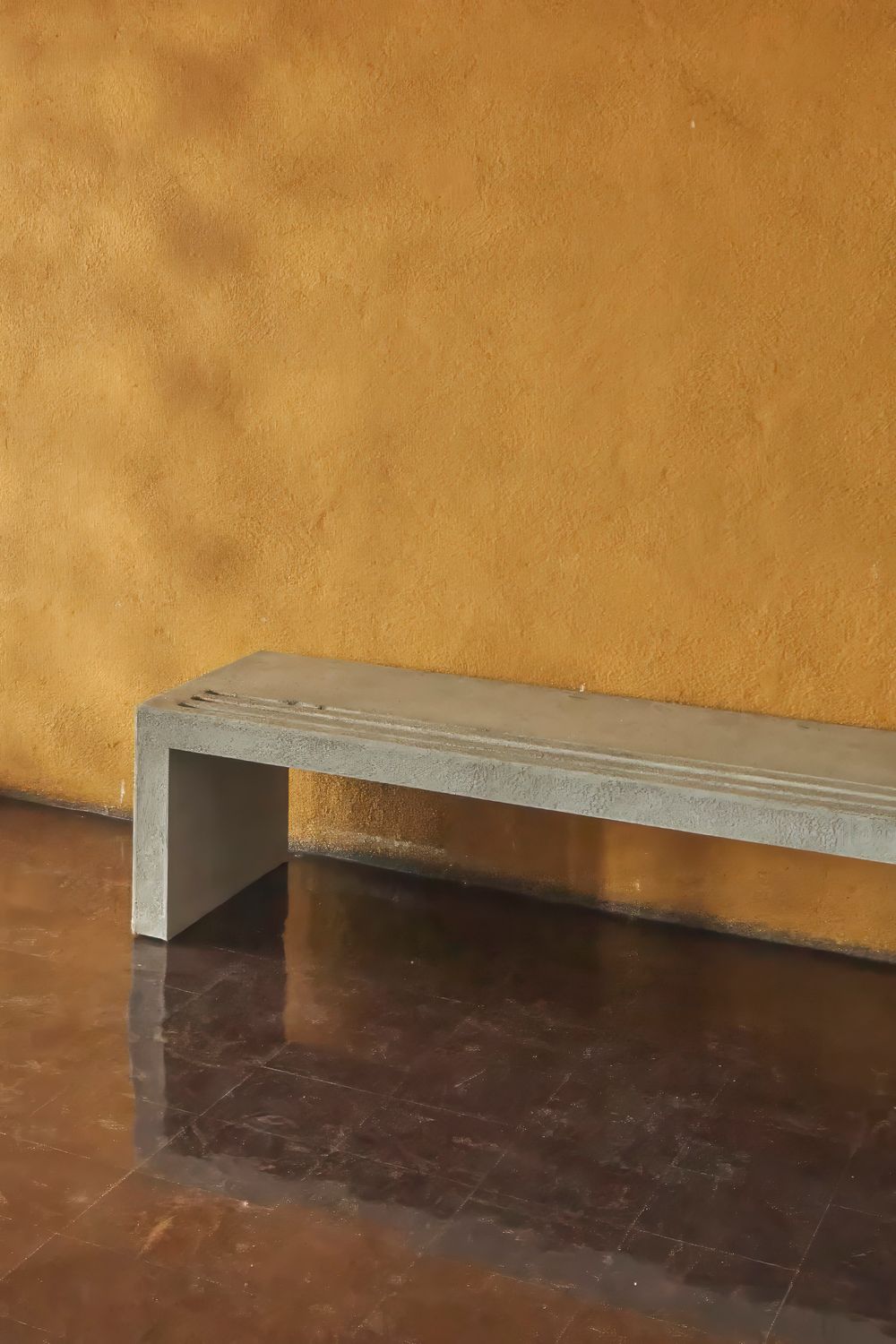 Minimalist concrete bench against a textured ochre wall on a glossy dark brown tiled floor in a warm, modern interior.