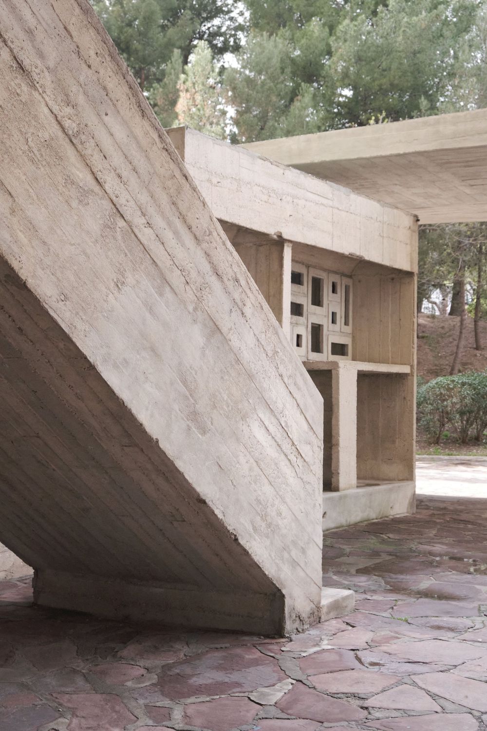 Board-formed concrete staircase and pavilion with a small perforated window panel, set on a flagstone terrace beneath pine trees.