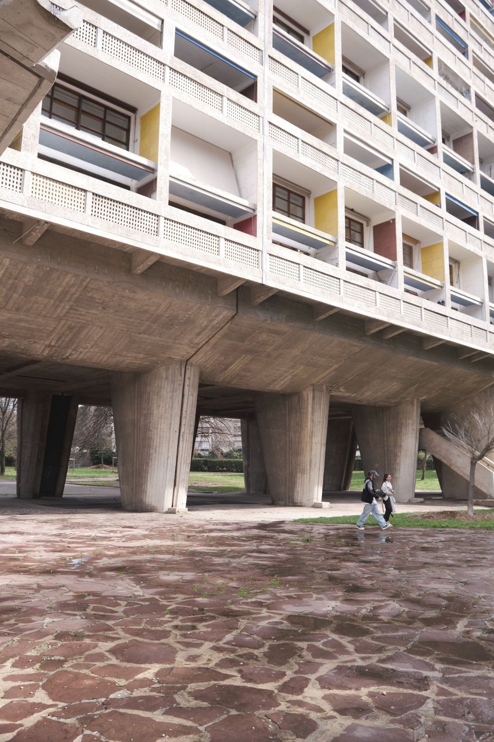 Brutalist concrete apartment block raised on massive pilotis with colorful balconies, as two people walk across the paved ground below.