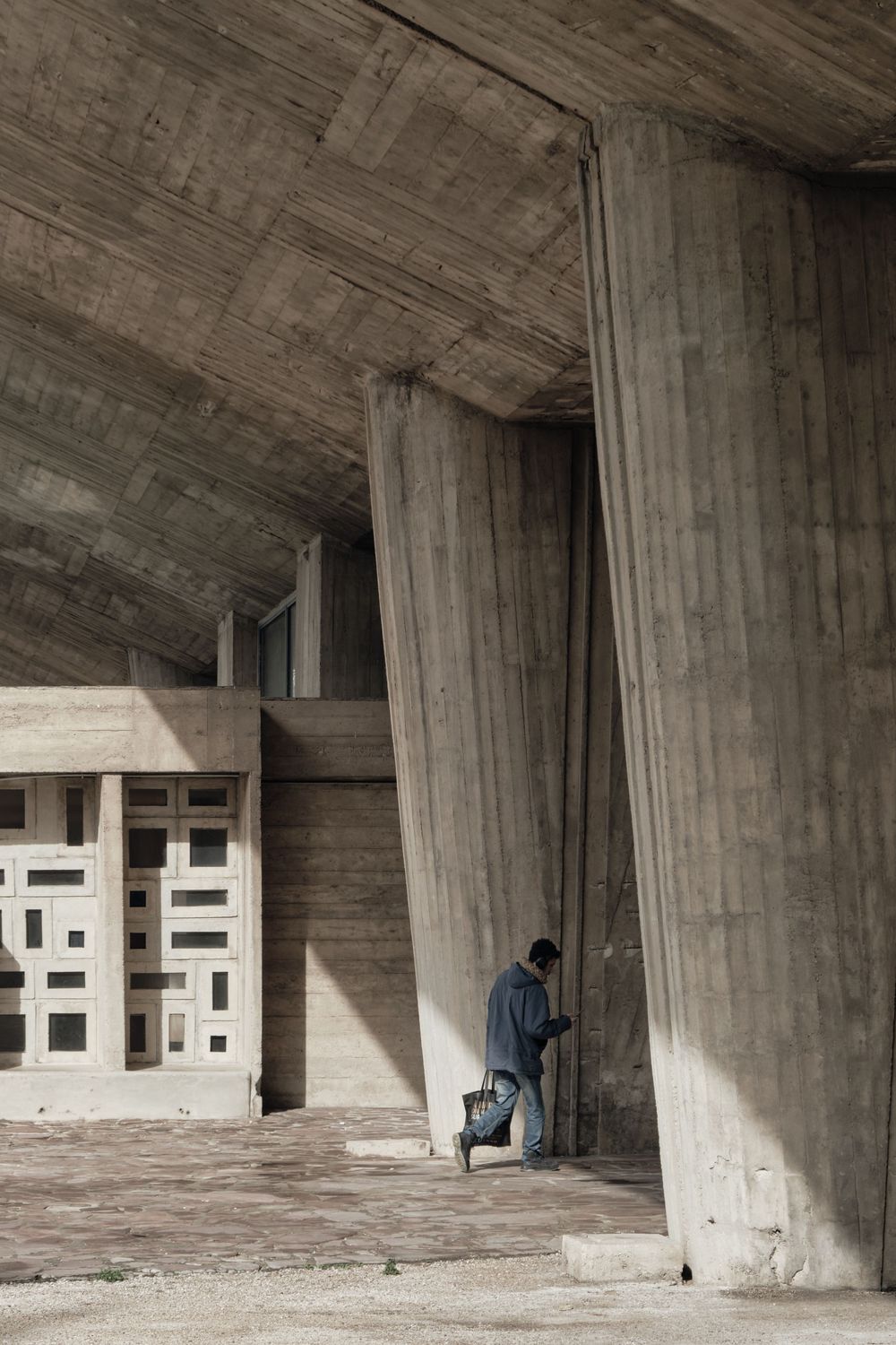 Man carrying a bag walks beneath massive board-formed concrete columns and a sweeping vaulted ceiling in brutalist architecture.