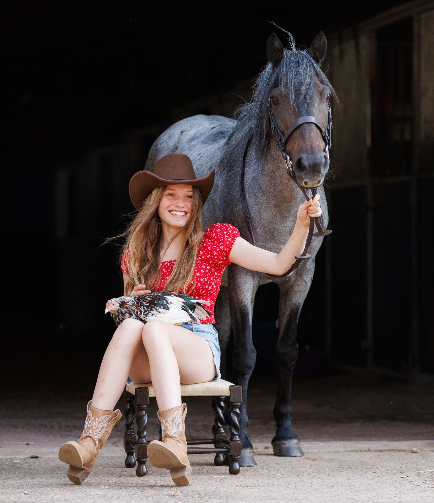 Smiling girl in a cowboy hat and boots sits on a wooden stool holding a chicken while a gray horse stands beside her in a dark barn.