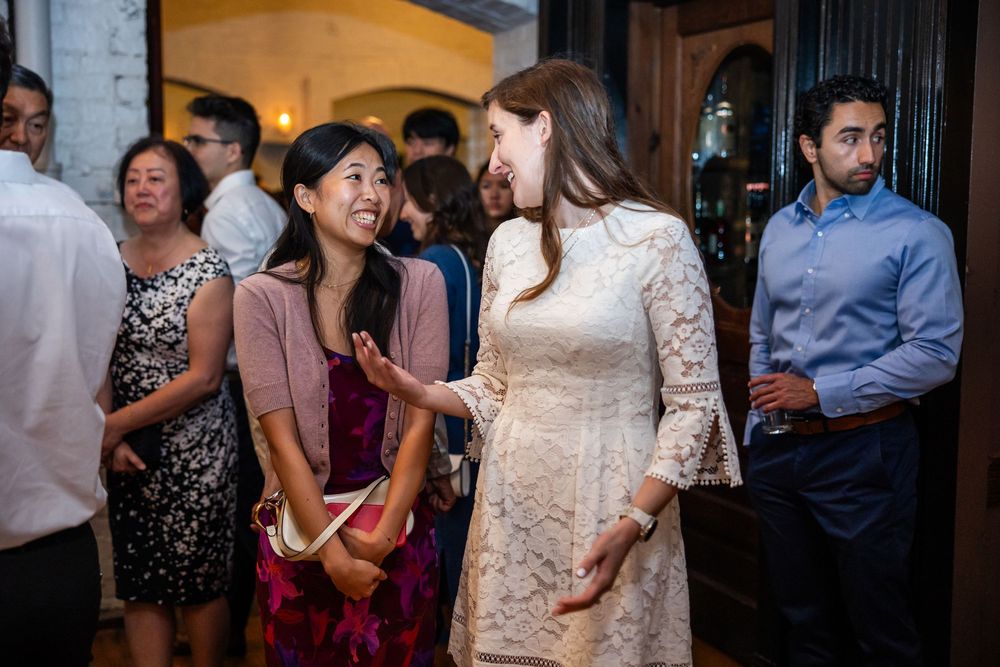 Two women in elegant dresses smile and chat at an indoor social gathering or wedding reception event.