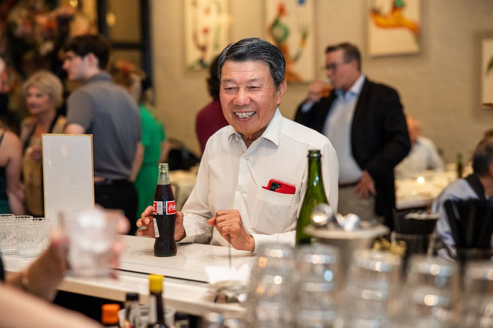 Smiling man holding a Coca-Cola bottle at a restaurant bar, enjoying a social gathering with guests in the background.