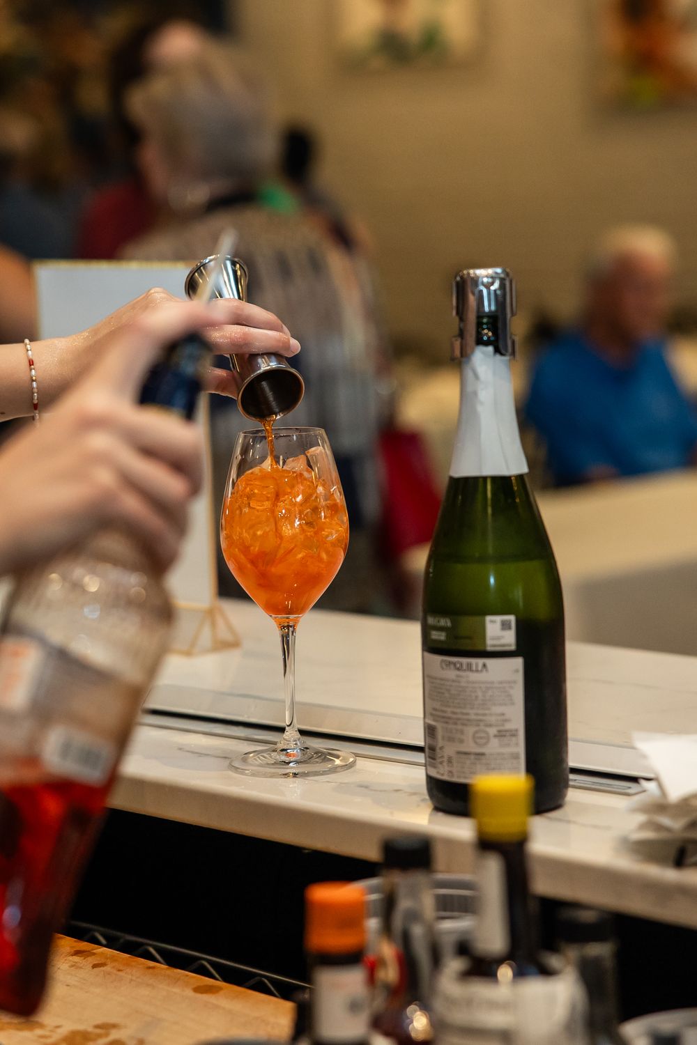 Bartender pouring a cocktail into a wine glass with ice and Aperol Spritz at a bar with a sparkling wine bottle.
