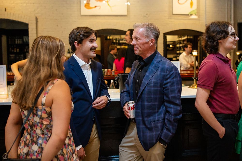 People networking at an indoor social event near a bar, engaged in conversation in a warmly lit venue.
