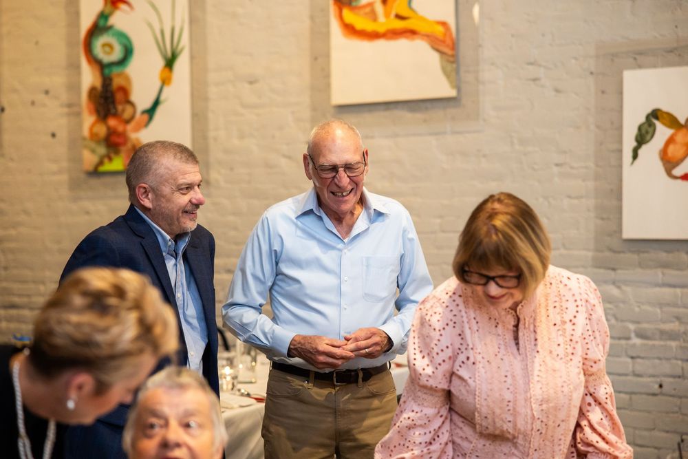 Three adults smiling and socializing at an art gallery event with colorful paintings displayed on brick walls.