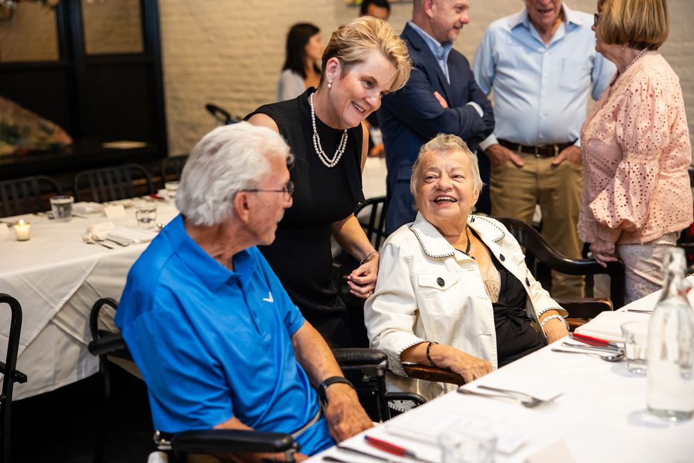 Elderly couple seated at a restaurant table smiling with a woman leaning in during a social gathering event.