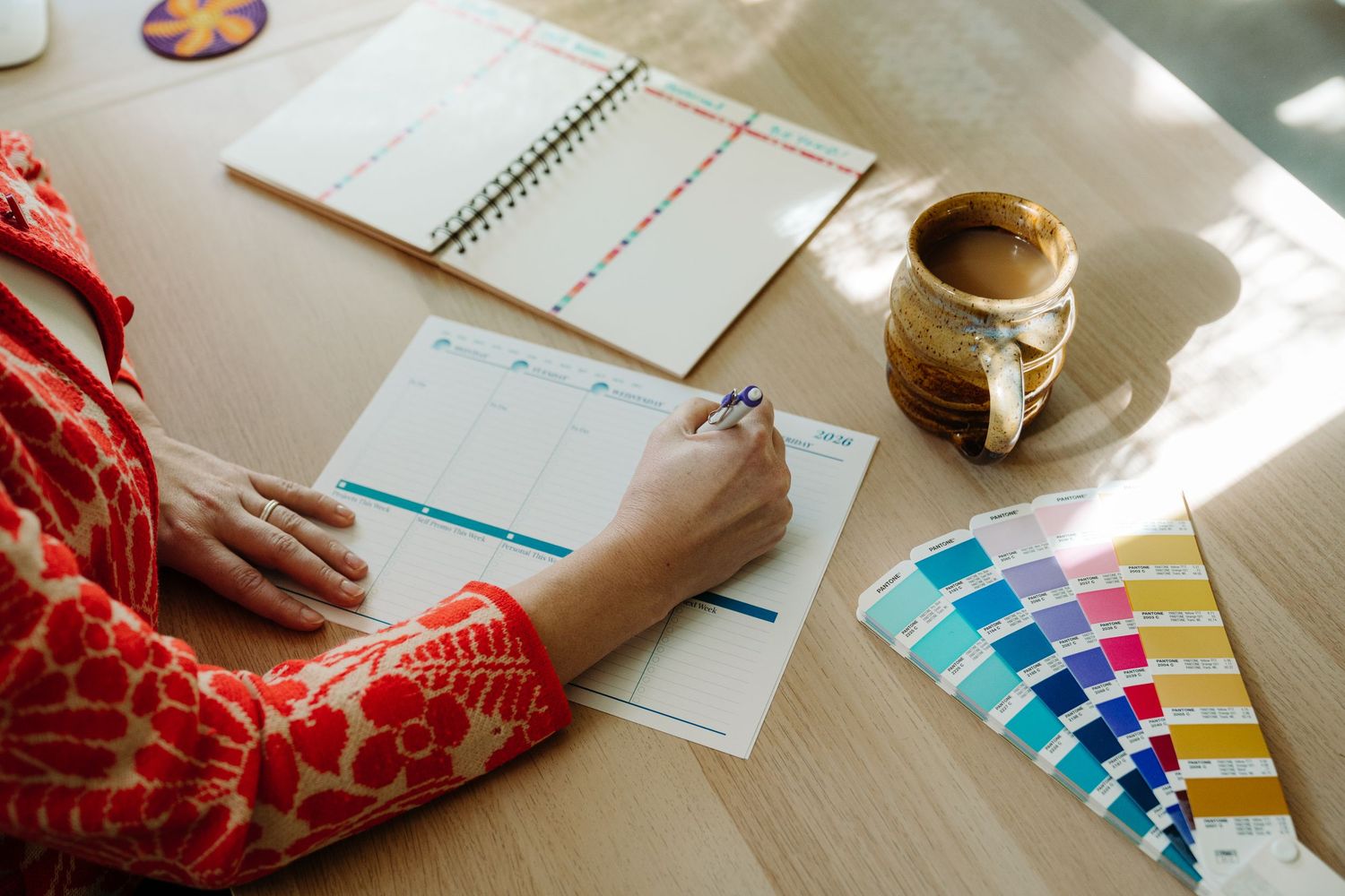 Designer reviewing color swatches and planning documents at a wooden desk with a coffee mug and open notebook.