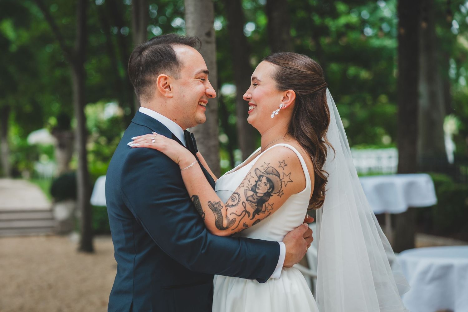 Smiling bride with tattoo and groom in navy suit embrace outdoors at wedding ceremony among trees outside the meridian house in dc
