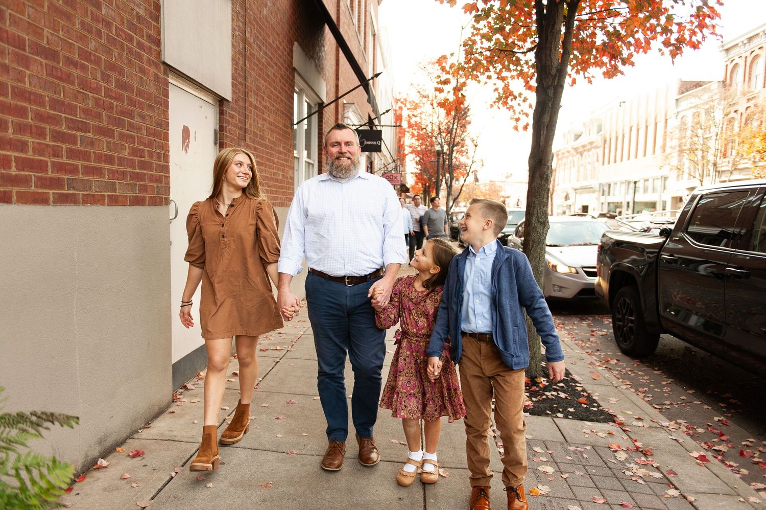 Family of four walking on a brick sidewalk in autumn, smiling and holding hands in a charming downtown setting.