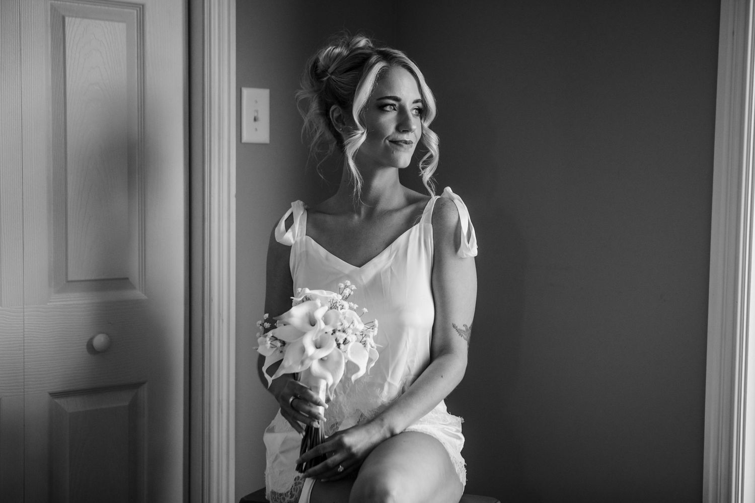 Black and white photo of a bride in a white dress holding a bouquet, seated by a door, gazing softly to the side.