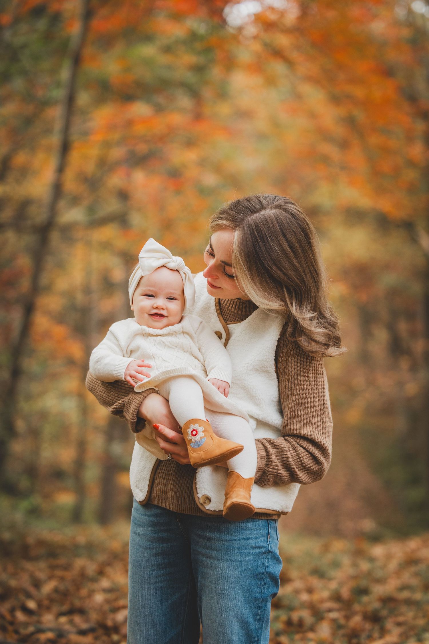 A young mother holds her smiling baby in a white bonnet and booties amid vibrant autumn forest foliage at Lake Fairfax Park