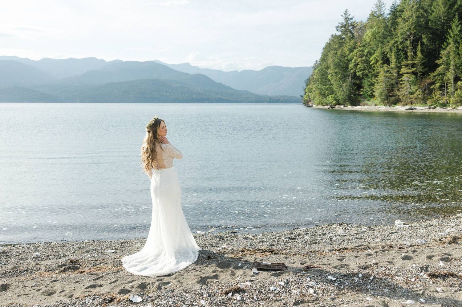 A bride and groom embrace on a rocky Pacific Northwest beach surrounded by driftwood, evergreen cliffs, and calm turquoise waters.