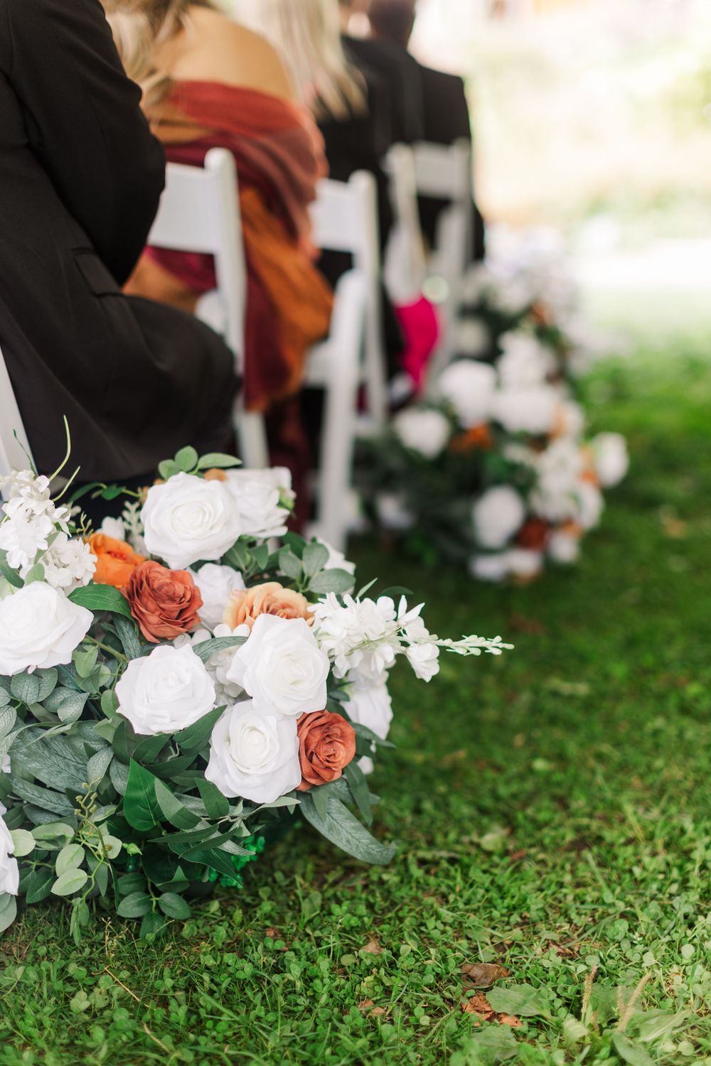 White and rust-orange rose arrangements with greenery line the grass aisle beside white chairs at an outdoor wedding ceremony.