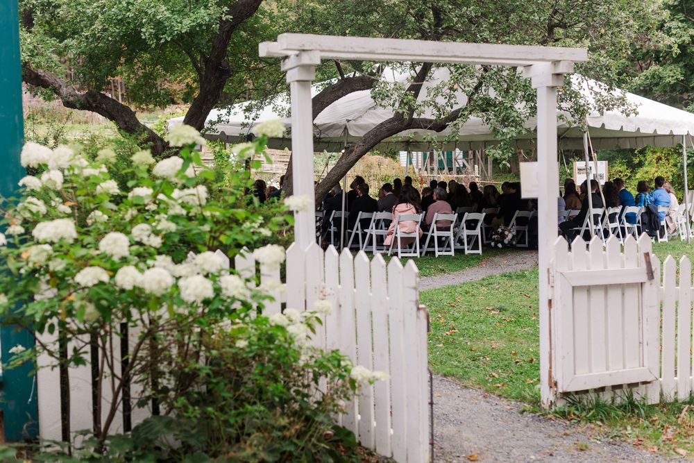 White picket fence and gate beside blooming white hydrangeas, with guests seated under a tent at an outdoor garden wedding ceremony.