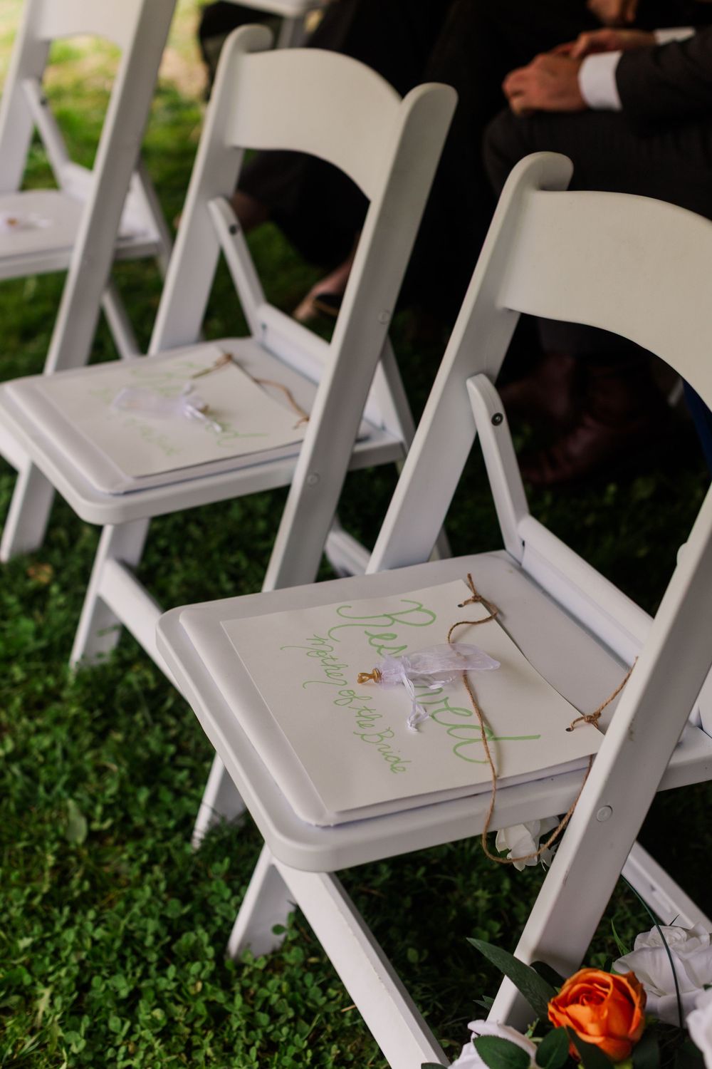White wooden folding chairs on green grass, each with a handwritten sign and ribbon on the seat, set up for an outdoor wedding ceremony.