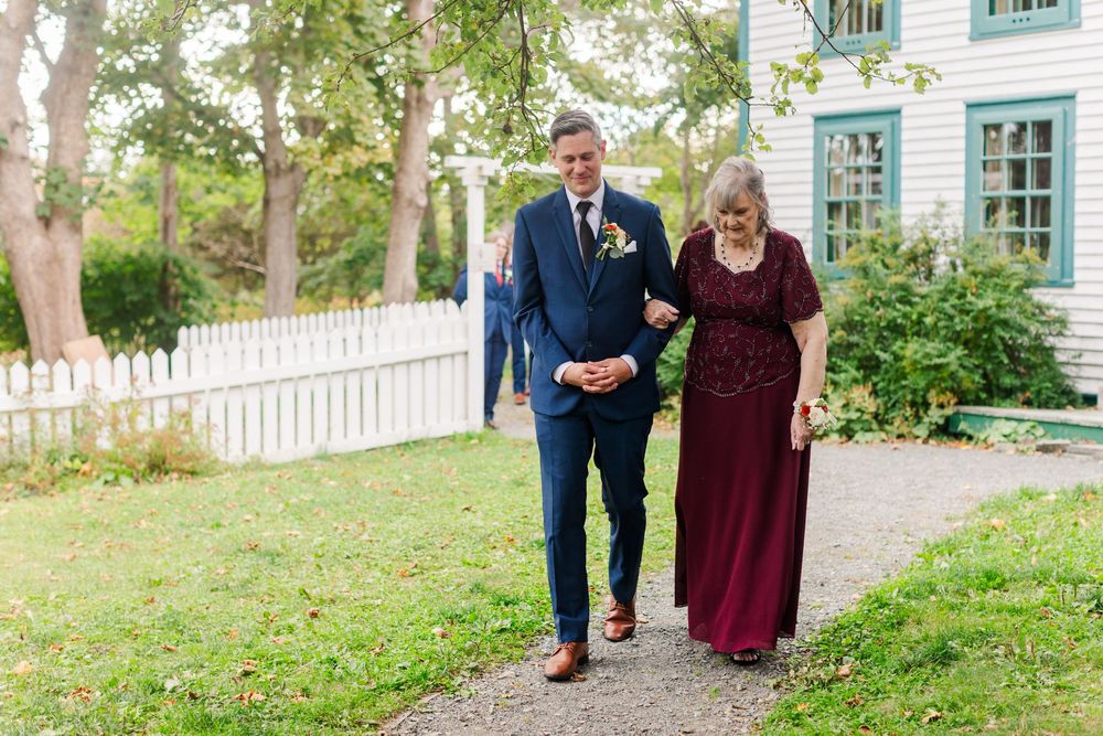 A groom in a navy suit escorts a woman in a burgundy lace dress along a garden path past a white picket fence at an outdoor wedding.