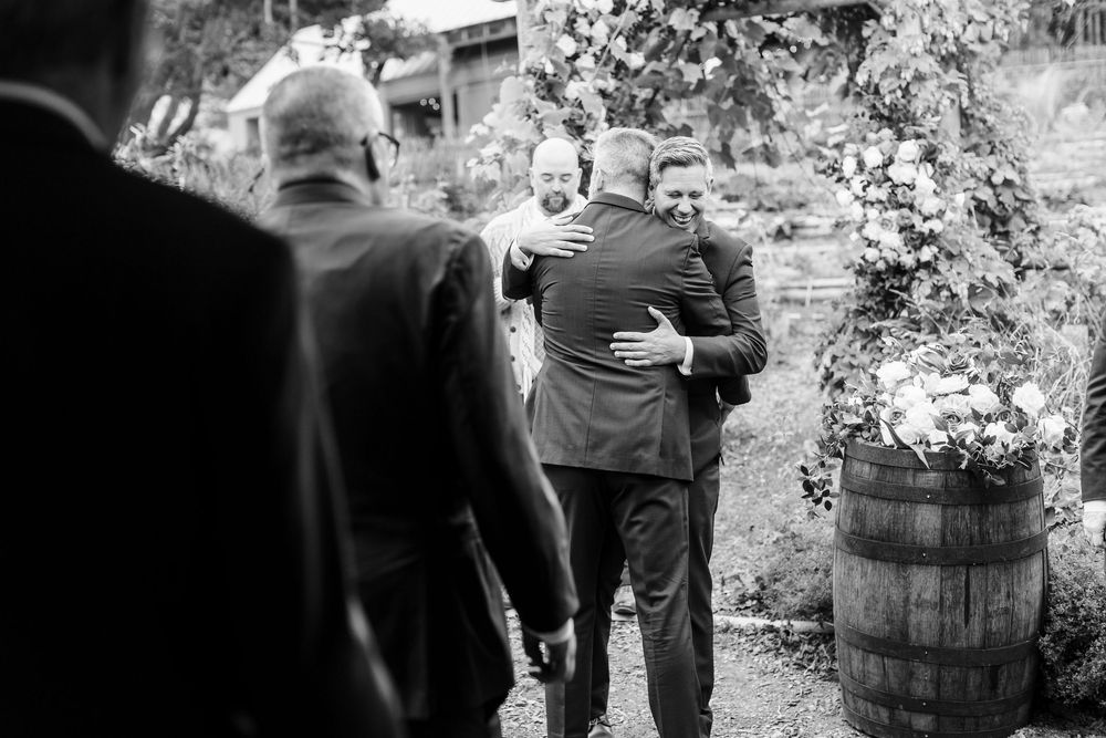 Two men in suits share a warm embrace at an outdoor wedding, with guests nearby and a floral arrangement on a wooden barrel.