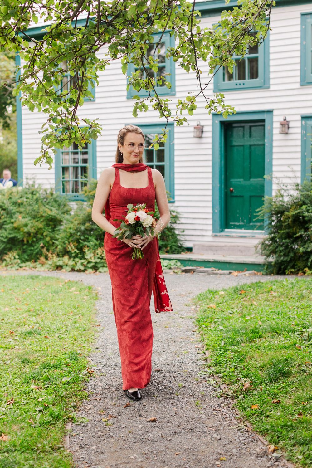 Woman in a red dress holds a bouquet of white and pink flowers while walking down a gravel path toward a white house with green trim.