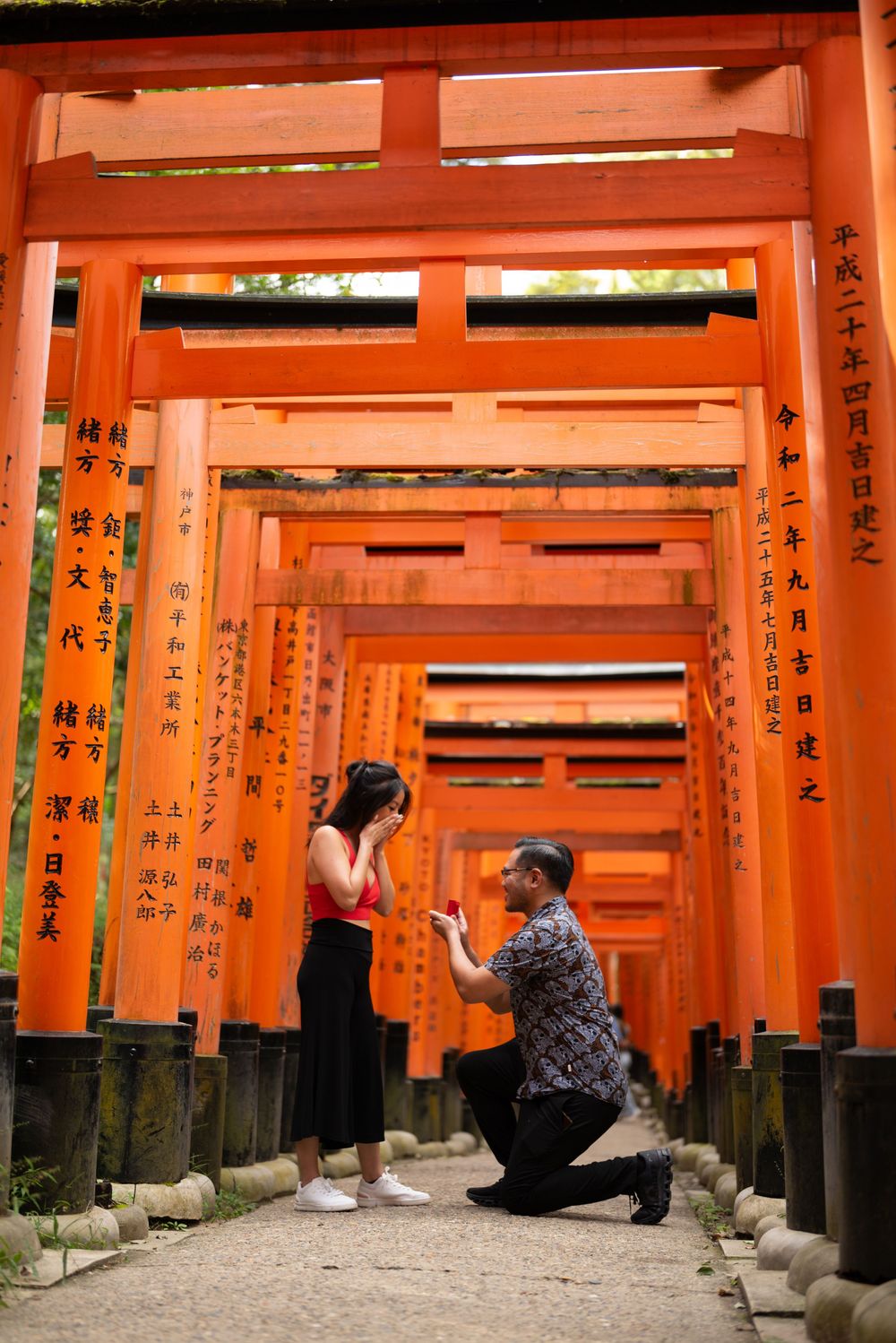 Man kneeling on one knee proposing to a woman covering her face among the orange torii gates at Fushimi Inari Shrine in Kyoto, Japan.