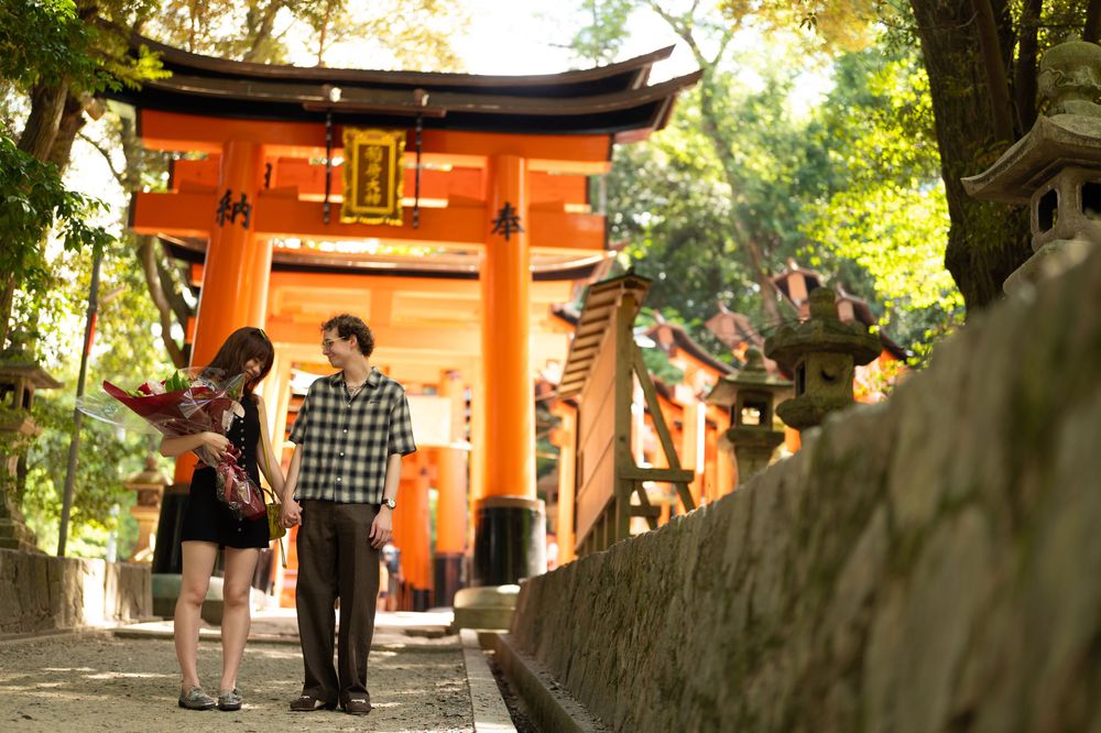Couple holding hands and walking toward a vibrant orange torii gate at a traditional Japanese shrine surrounded by green trees.
