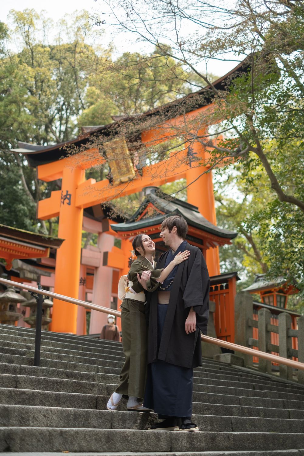 Couple in traditional Japanese attire embracing on stone steps beneath a vermilion torii gate at a Kyoto shrine surrounded by trees.