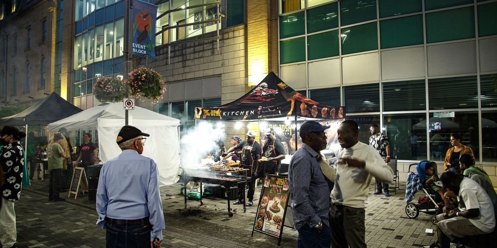 Nighttime outdoor street food market with vendors and shoppers gathered under bright lights near a modern building.