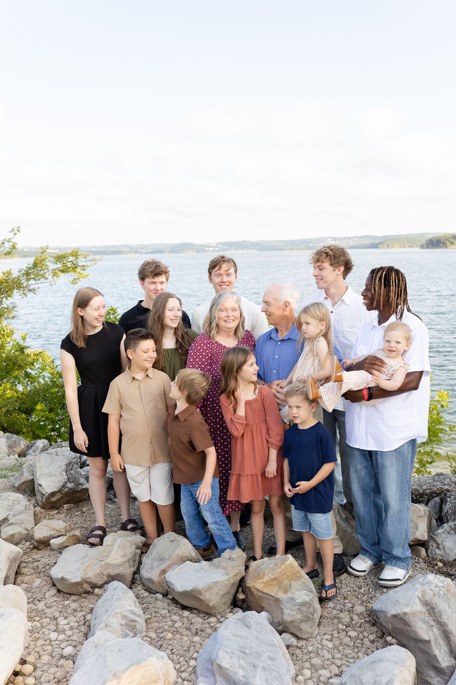 Large multigenerational family posing together on rocky lakeside shore surrounded by lush greenery on a bright summer day.
