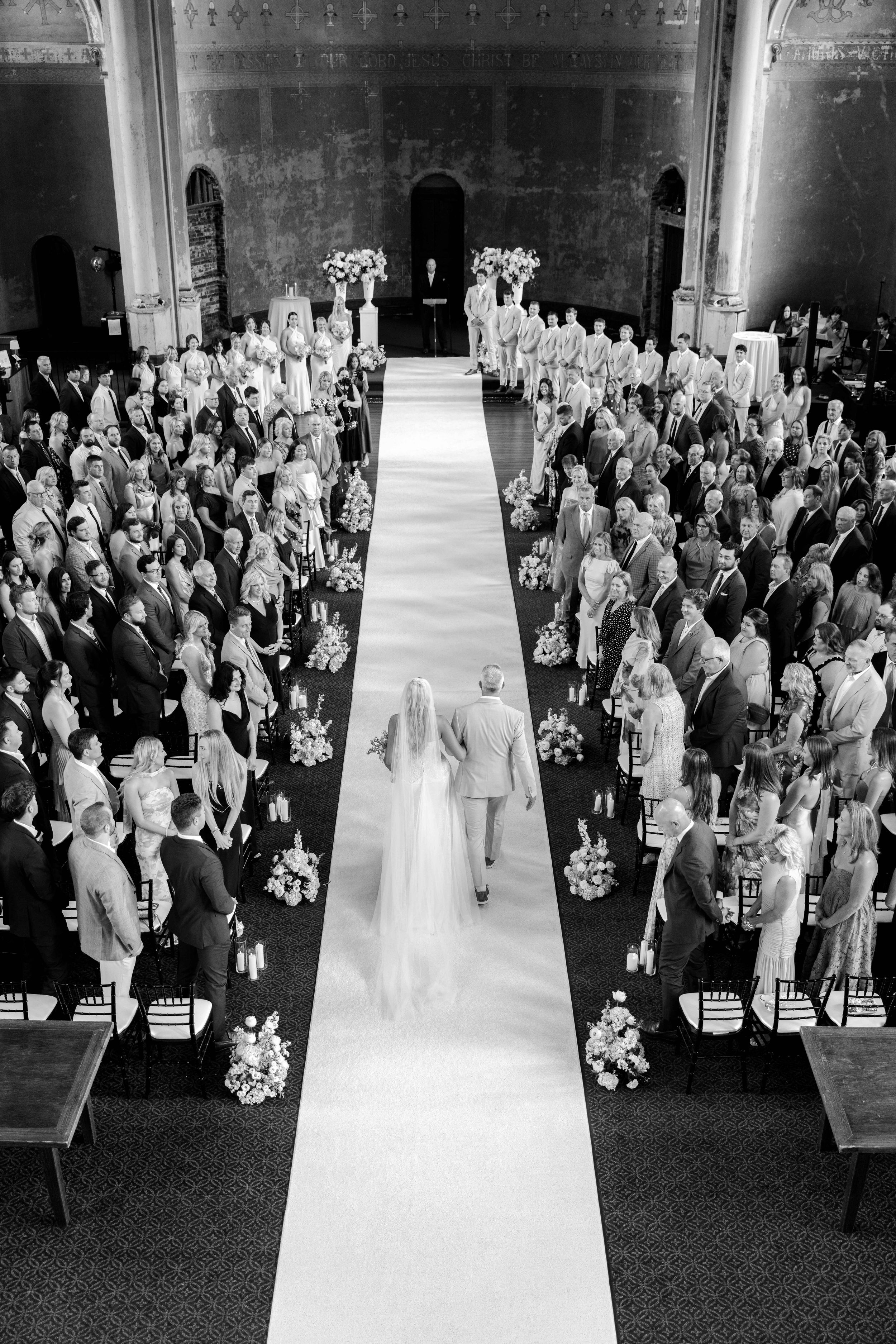 Aerial black and white photo of bride walking down the aisle at an elegant wedding ceremony with guests standing.