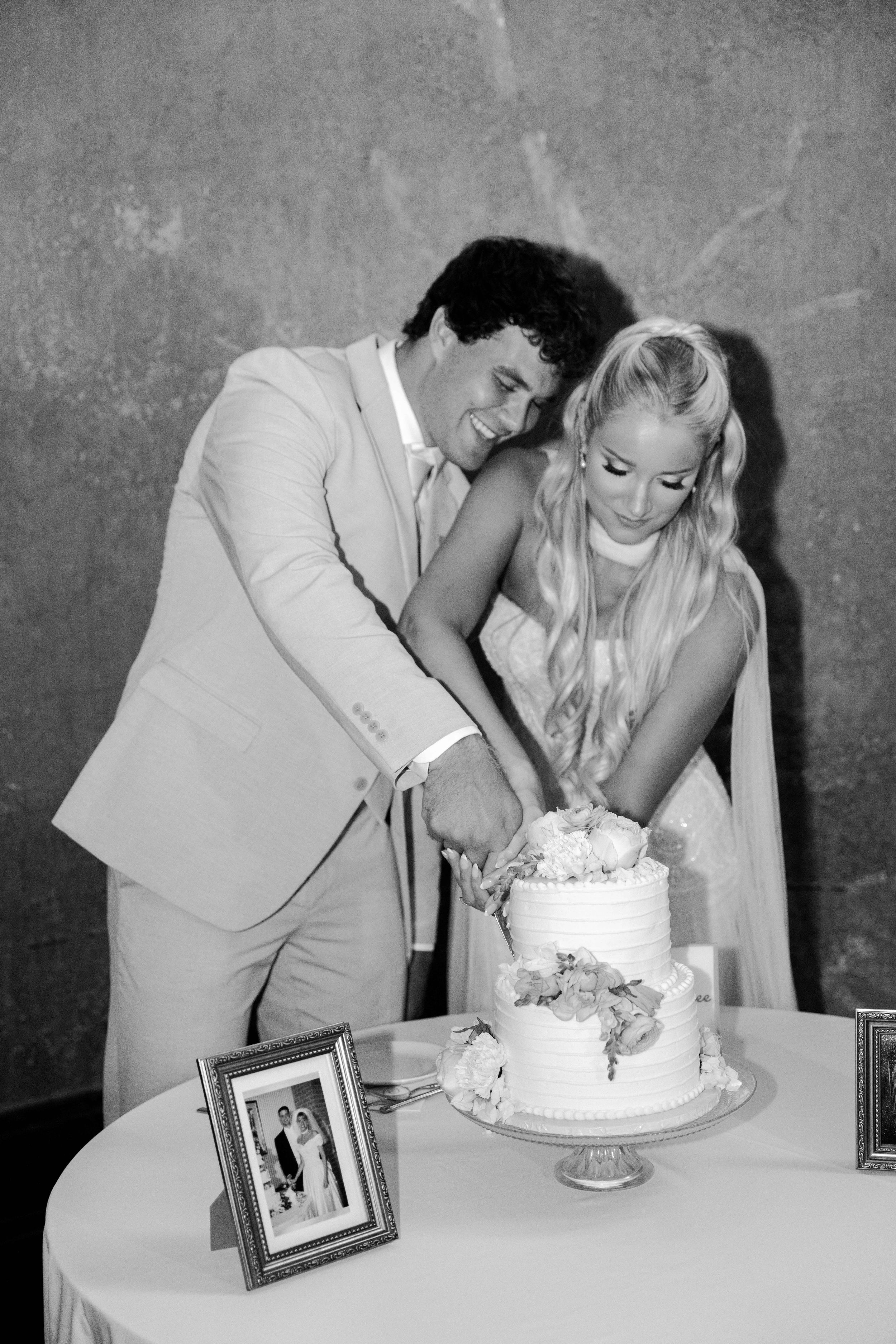 Black and white photo of bride and groom cutting wedding cake together, smiling joyfully at reception.