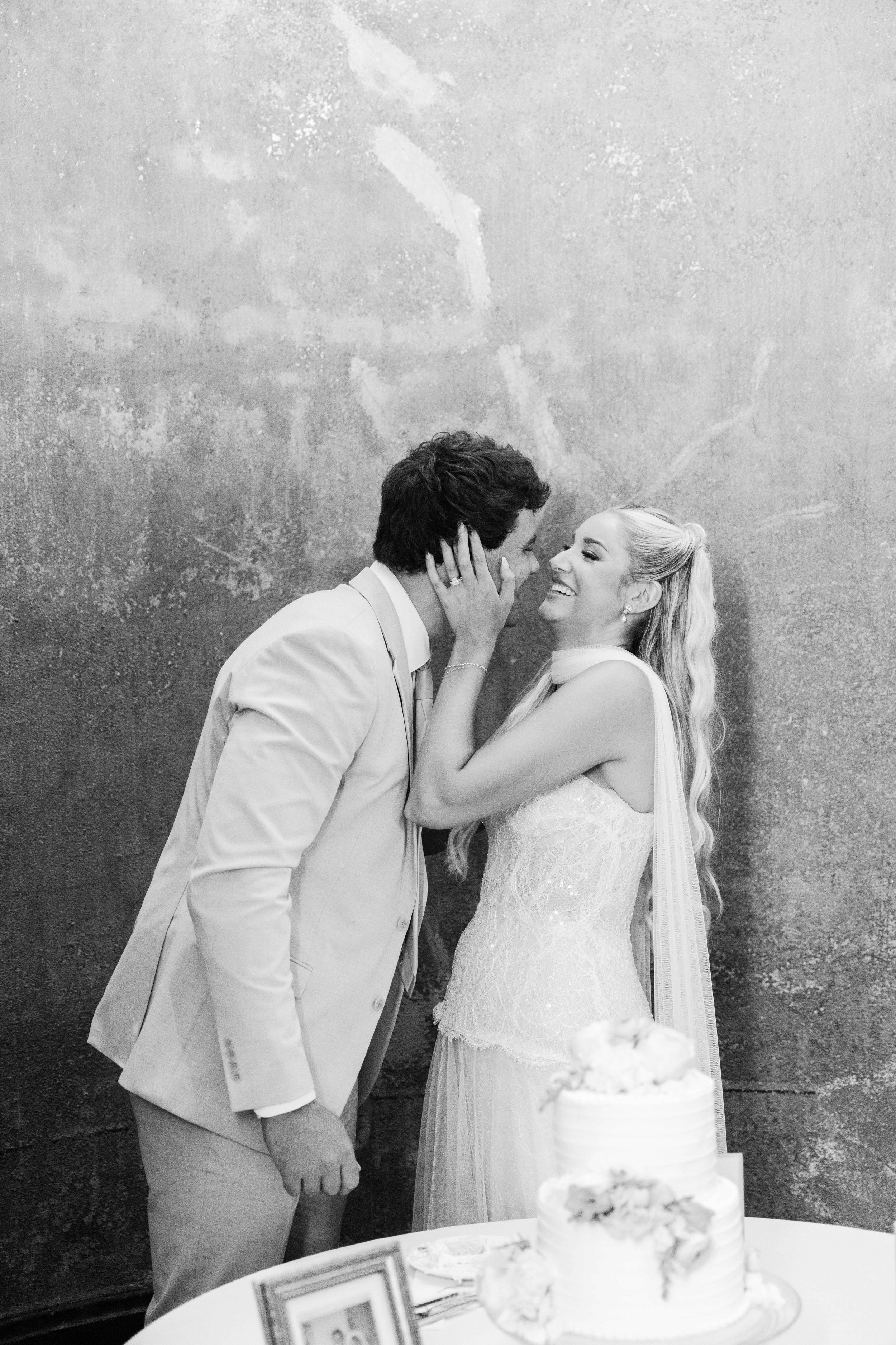 Black and white photo of a smiling bride and groom sharing a kiss near their wedding cake.