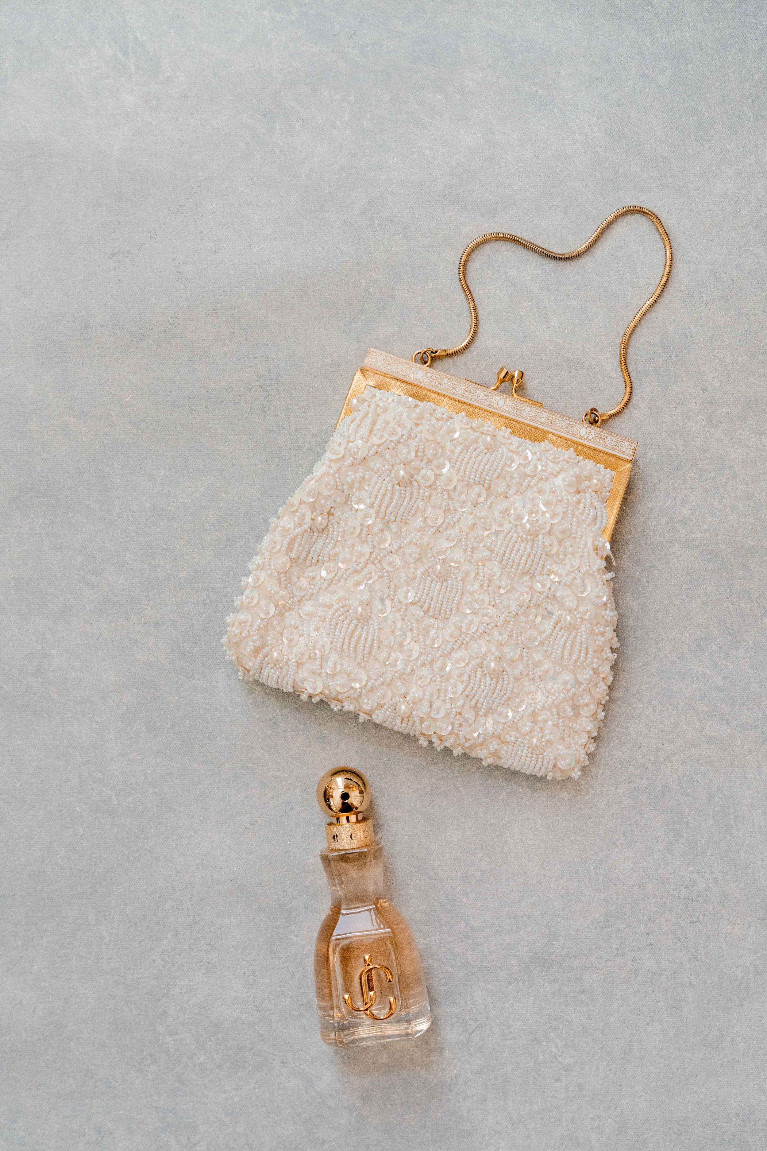Elegant white beaded bridal clutch purse with gold frame handle next to a small perfume bottle on gray background.