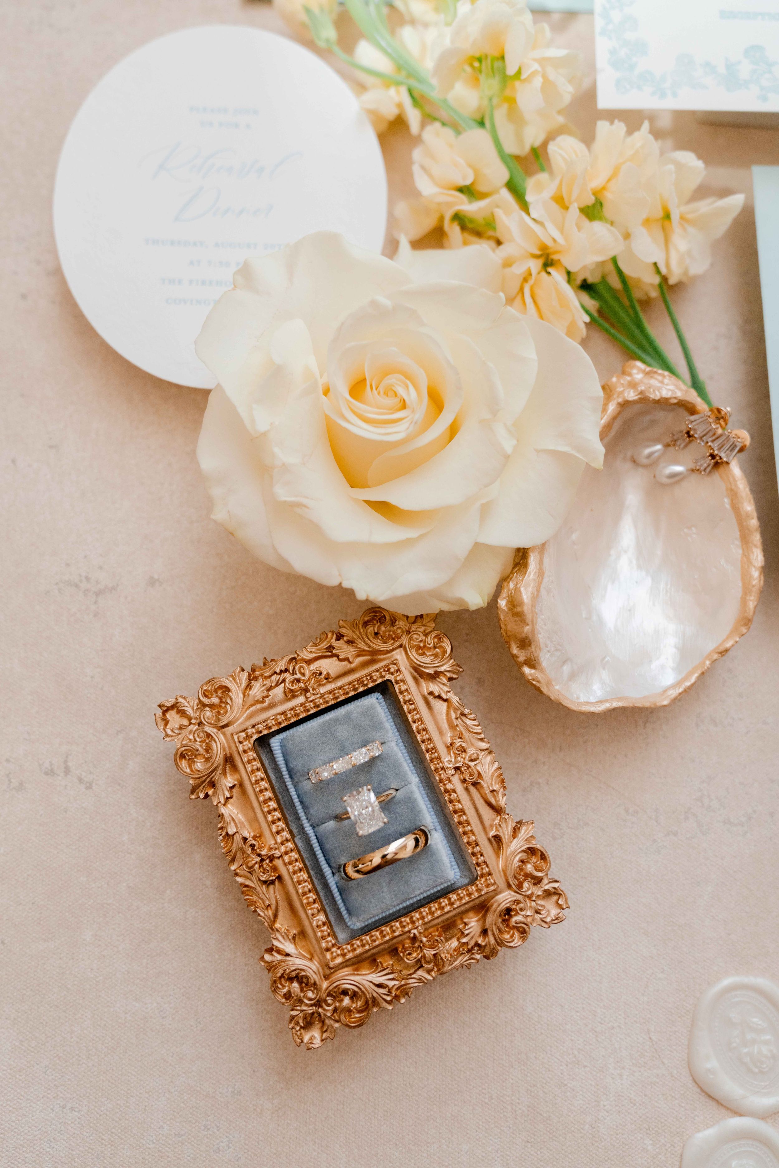 Elegant wedding rings displayed in ornate gold frame with white rose and floral arrangement.