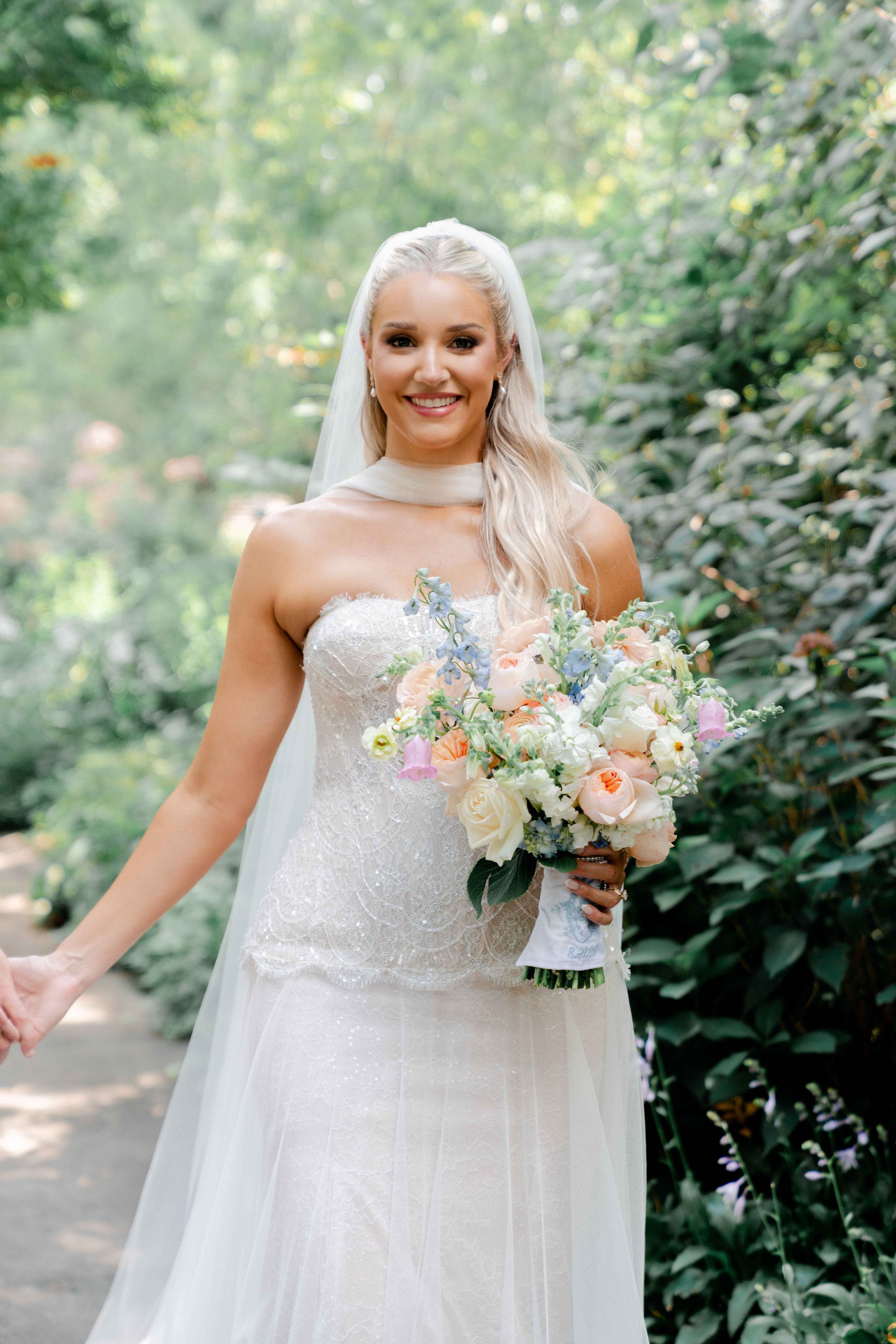 Smiling bride in strapless lace gown holding pastel bouquet of roses and blue flowers in a lush garden setting.