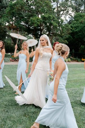 Blonde bride in white lace wedding gown walks with bridesmaids in light blue dresses carrying parasols outdoors.