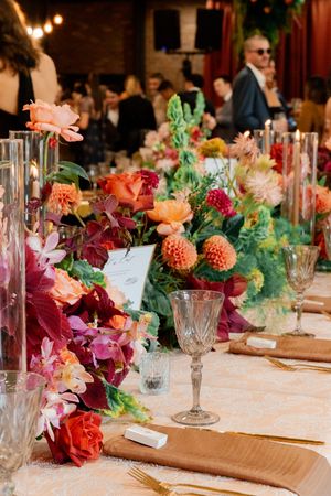 Elegant wedding reception table with vibrant floral centerpieces in orange, pink, and red, crystal glassware, and gold cutlery.