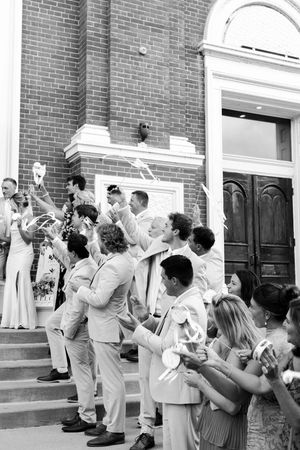 Black and white photo of wedding party celebrating on church steps, waving ribbon wands as couple kisses.