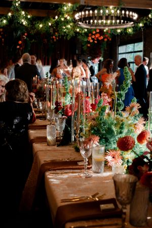 Elegant wedding reception table with colorful floral arrangements, candles, and guests mingling under chandelier lighting.