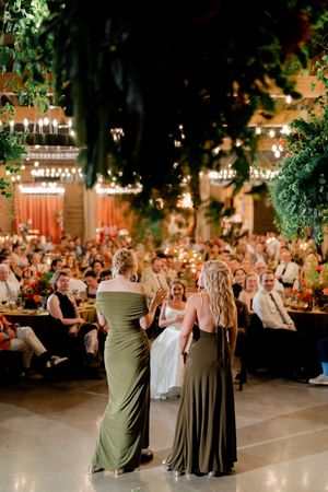 Two bridesmaids in olive green gowns give a speech at an elegantly decorated wedding reception with string lights and lush greenery.