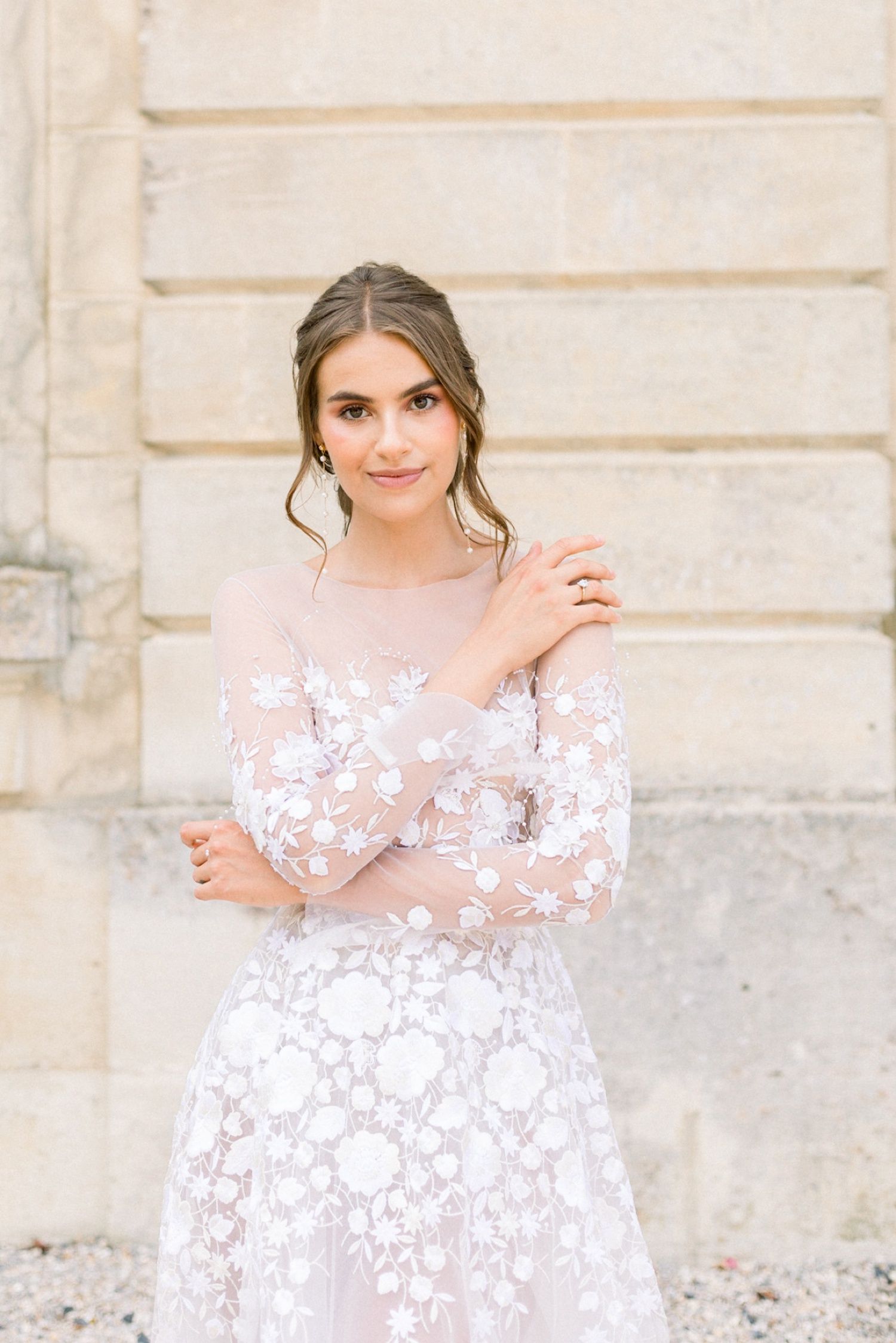 Elegant bride in sheer floral lace long-sleeve wedding dress posing before a stone building.
