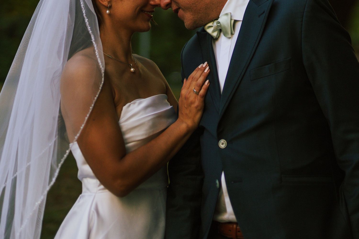 Bride in white gown with veil and groom in dark suit walking on grass near trees and building.
