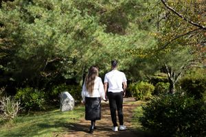 Couple in white shirts walking hand in hand along a dirt path through a peaceful, lush green garden with trees and shrubs.