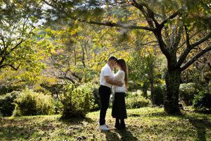 Couple in white shirts and dark bottoms embracing on a sunlit grassy path surrounded by tall trees and lush green foliage in a park.