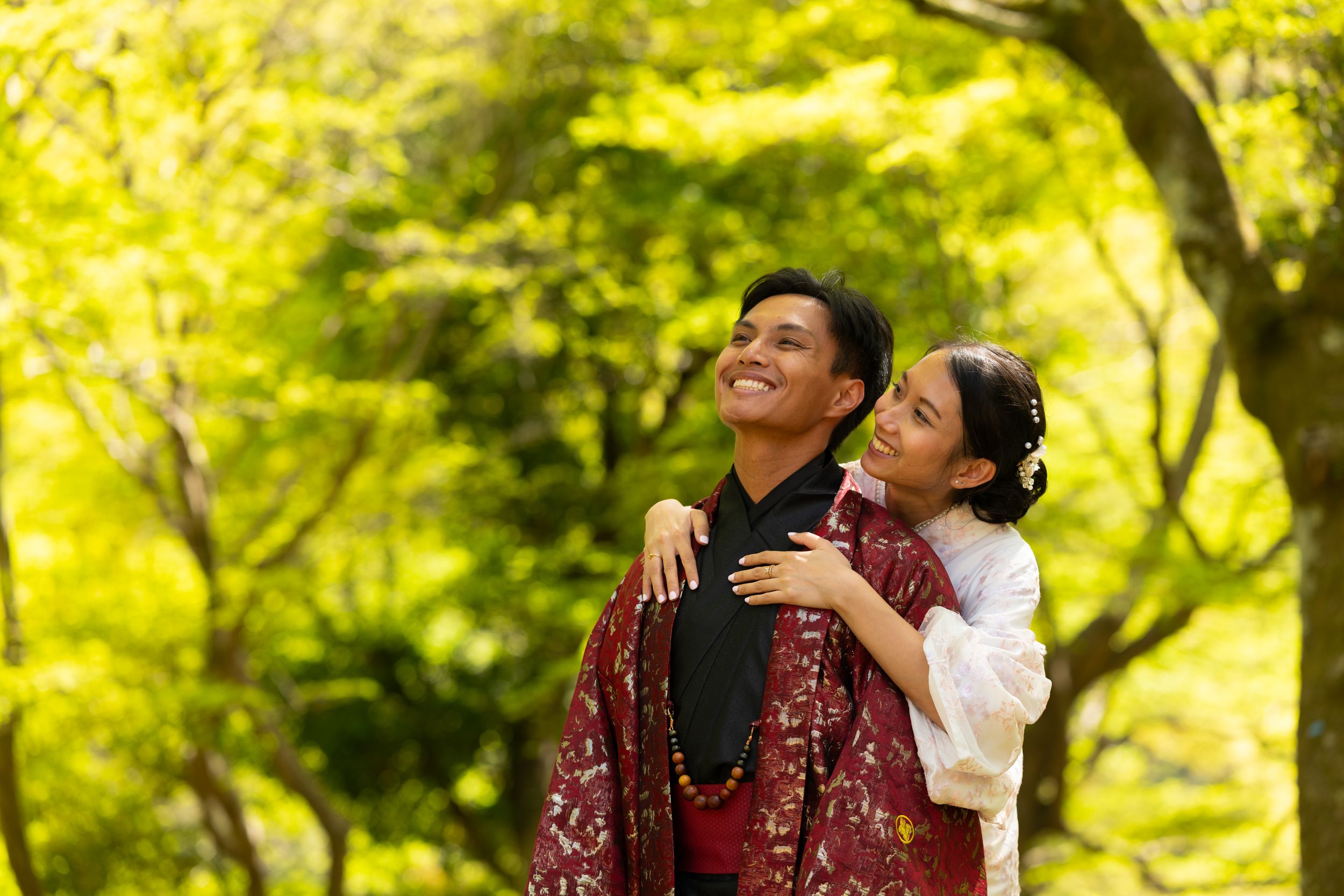 A smiling couple in traditional Japanese kimono embrace outdoors, with the woman hugging the man from behind among bright green trees.
