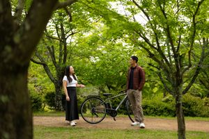 A young woman and man stand beside a parked bicycle on a dirt path surrounded by lush green trees in a sunlit park.