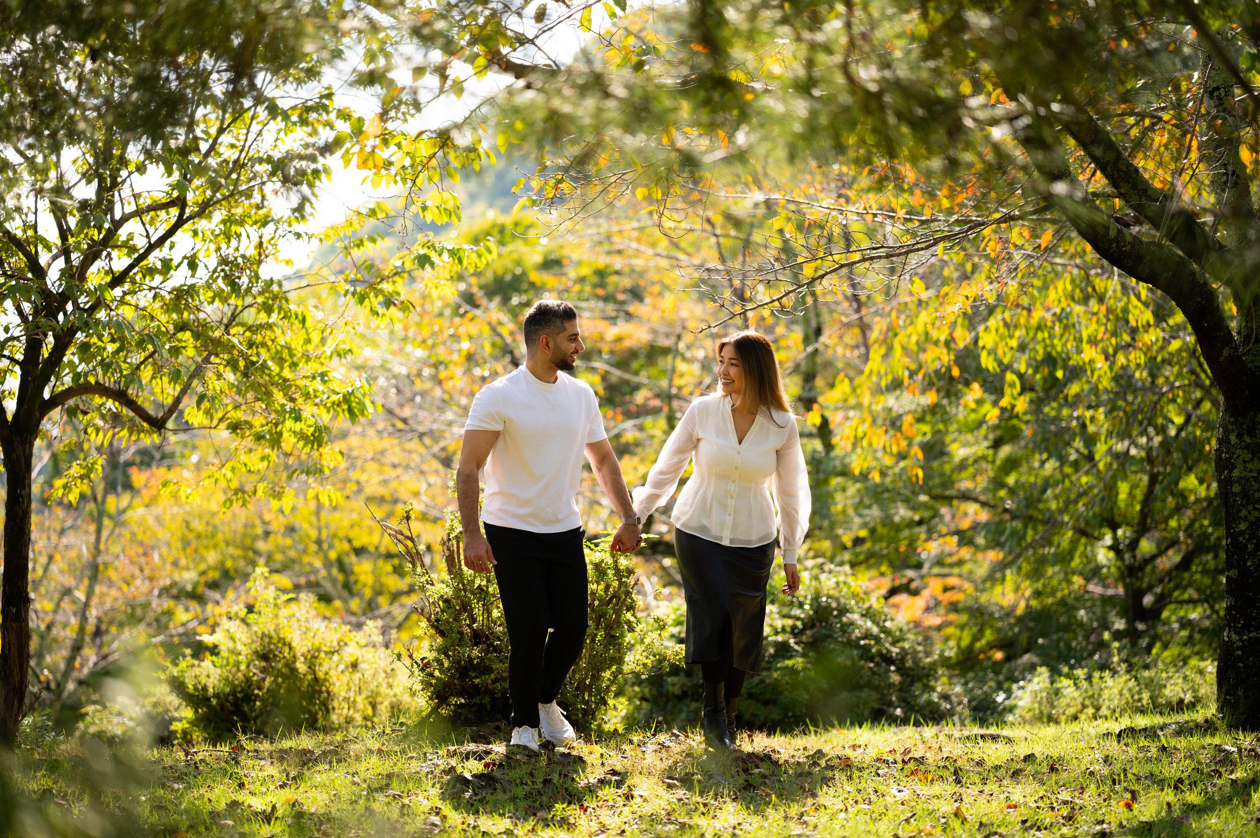 Couple in casual clothes holding hands and walking through a sunlit park surrounded by green trees and autumn foliage.