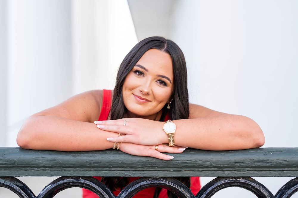 Smiling young woman with long dark hair in a red top rests her arms on a black wrought iron railing, wearing a gold watch.