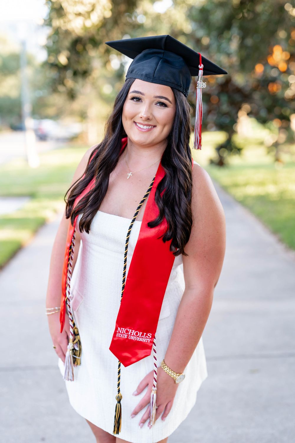 Smiling Nicholls State University graduate in a black cap, red stole, and graduation cords poses in a white dress on a tree-lined sidewalk.
